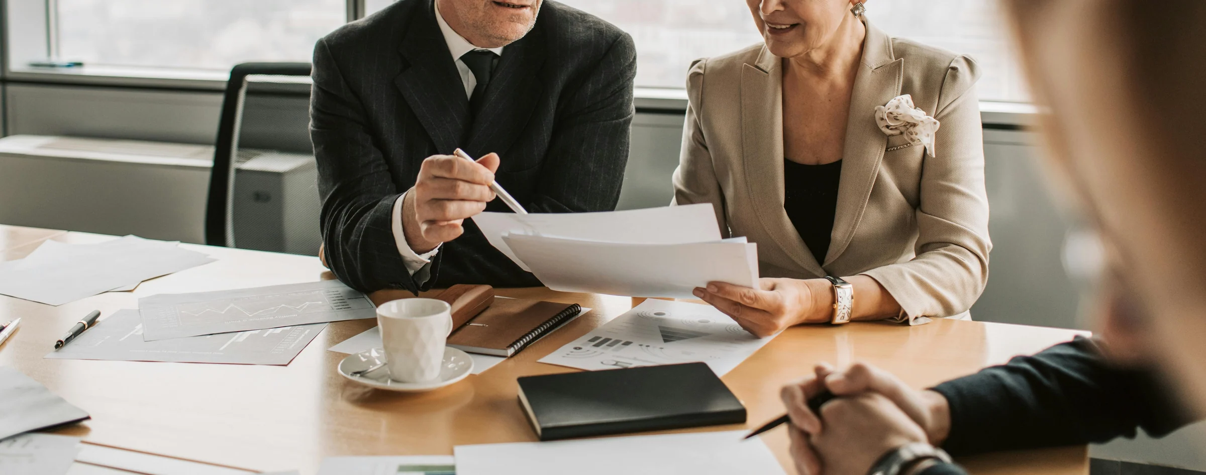 Duas executivas analisando documentos de planejamento financeiro sobre a mesa, diante das janelas de um escritório.